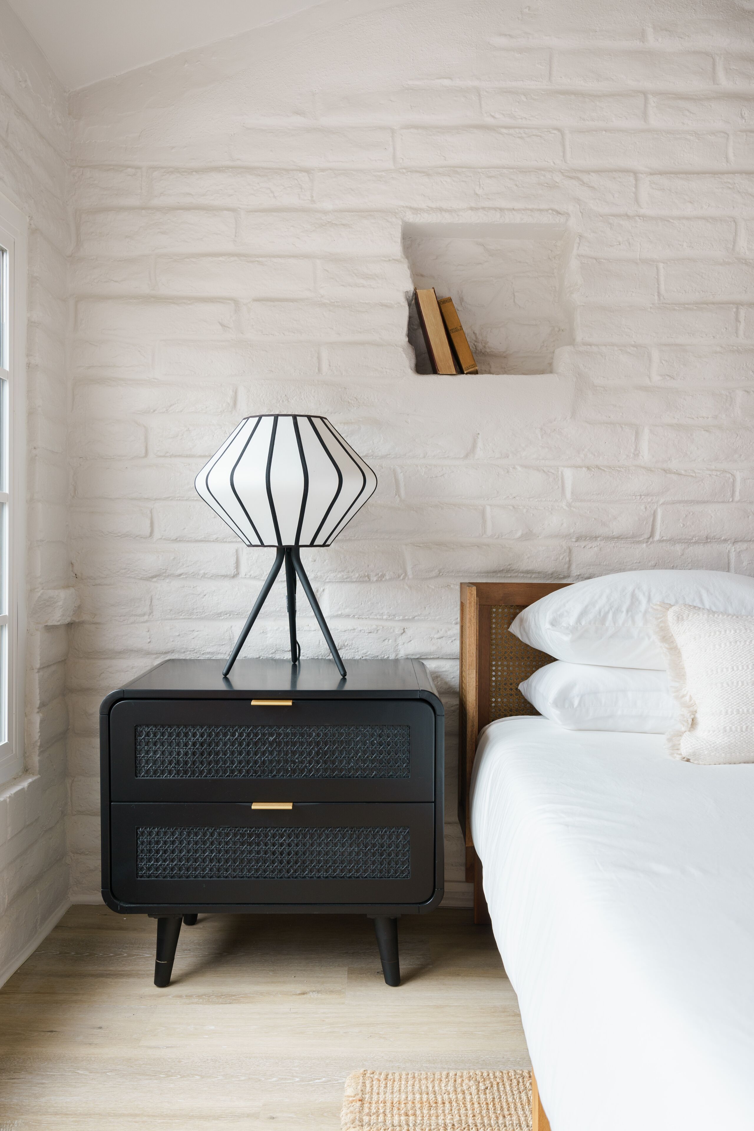 Quarto com parede de tijolos brancos, mesa de cabeceira preta com abajur, cabeceira de madeira e cama com roupa de cama branca.