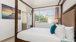 Ein Schlafzimmer mit einem hölzernen Himmelbett, einem großen Fenster mit Jalousien und einem Gemälde, das einen Sonnenuntergang über Wasser zeigt.