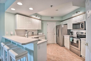 A kitchen with white cabinets, a stainless steel refrigerator, a microwave, an oven, a sink, and a countertop with stools. - Walk to Ocean: Condo w/ Pool in New Smyrna Beach (New Smyrna Beach)