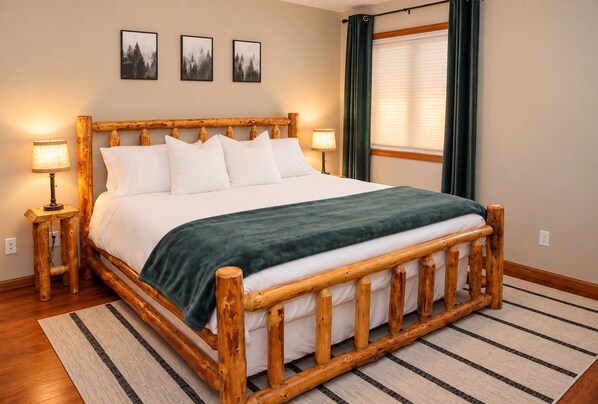 Una cama con estructura de madera, ropa de cama blanca y una manta verde, flanqueada por dos lámparas de noche, en una habitación con un tapete a rayas y cuadros en la pared.
