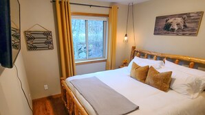 A bedroom with a wooden bed frame, white bedding, a window with curtains, a wall-mounted light fixture, and a painting. - Cozy pet-friendly home near hiking trails and Lake Vermilion (Tower)