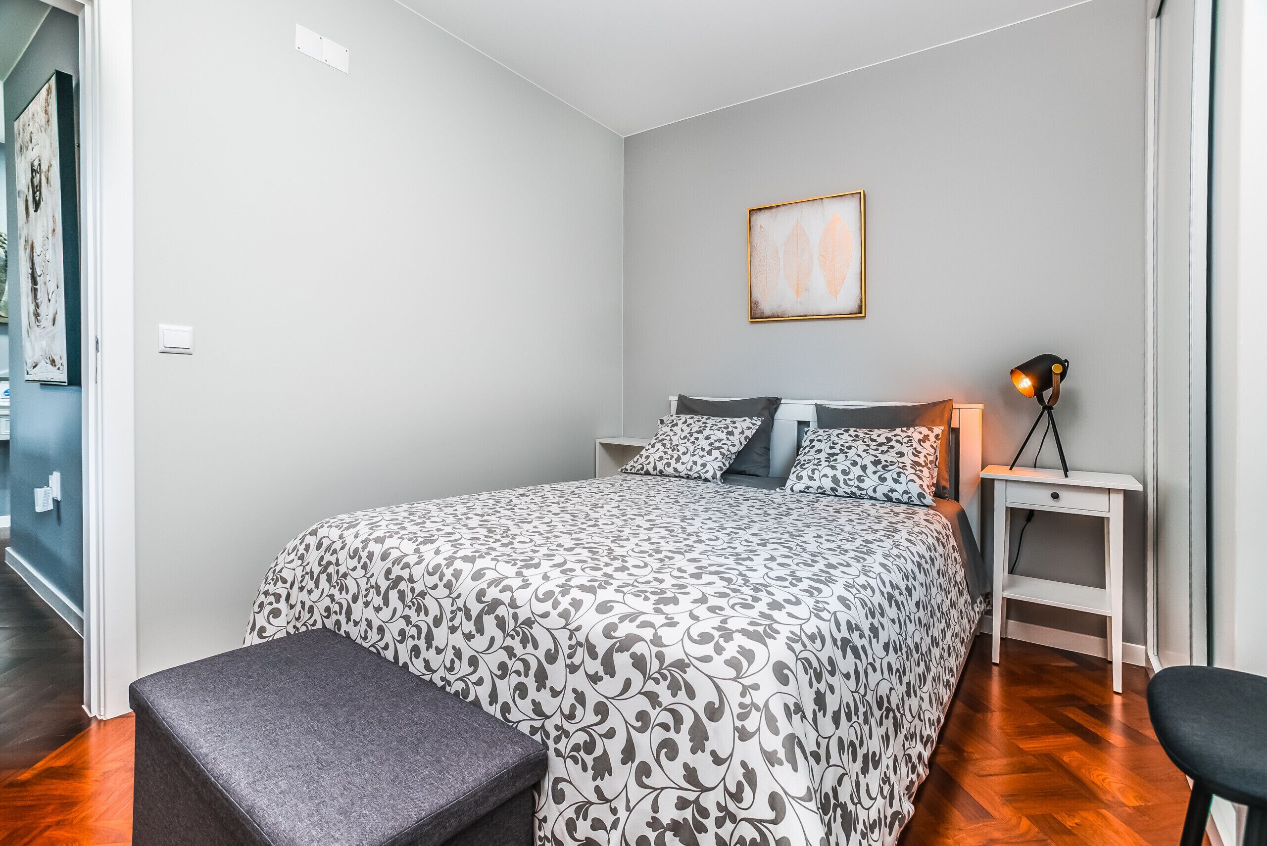 A bedroom with a bed, a bedside table with a lamp, a bench, and a framed picture on the wall.