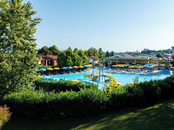 A swimming pool with a water slide, surrounded by lounge chairs and umbrellas, with buildings and trees in the background.