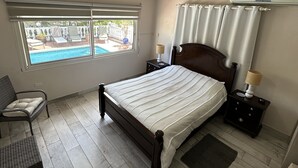 A bedroom with a large bed, a window with a view of a swimming pool, a ceiling fan, and a nightstand with a lamp. - Villa Belfiore (Collectivity of Saint Martin)