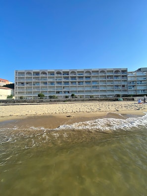 Un hôtel à plusieurs étages, doté de balcons, situé près d’une plage de sable où l’on peut observer les vagues.
