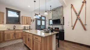 A modern kitchen with wooden cabinets, a granite countertop, pendant lighting, a stone fireplace, and skis mounted on the wall. - Brand New Condo Near Winter Park - Free Shuttle To Ski Resort- Private Hot Tub (Fraser)