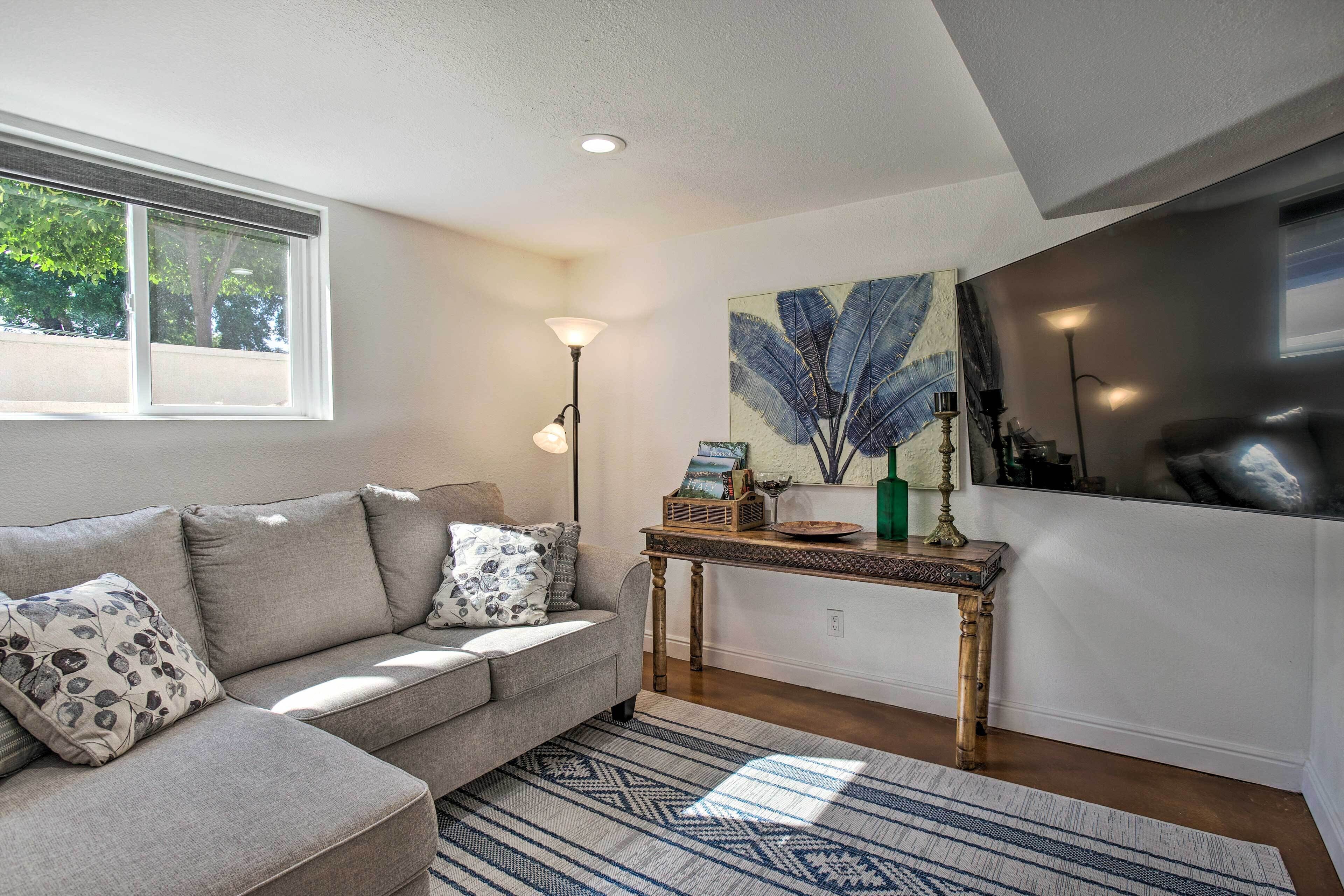 A living room with a grey sofa, patterned cushions, a wooden side table with decorative items, a large abstract painting, and a window with a view of trees.