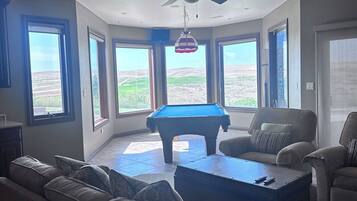 A living room with a pool table, a ceiling fan, and large windows offering a view of the outdoors.