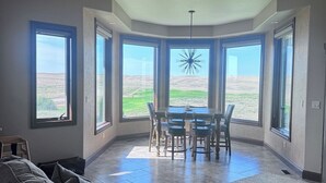 A spacious living room with a large gray sofa, a dining area with a table and chairs, and large windows offering a view of the outdoors.