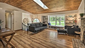 A spacious living room with wooden flooring, a black sofa, a dark armchair, and a built-in bookshelf. - Lake Nagawicka Home - 2 Blocks to Boat Launch (Delafield)
