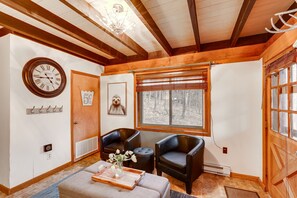 Ein gemütliches Zimmer mit Holzboden, einem großen Fenster mit Jalousien, zwei schwarzen Ledersesseln, einem Holztisch mit Tablett und einer Wanduhr.