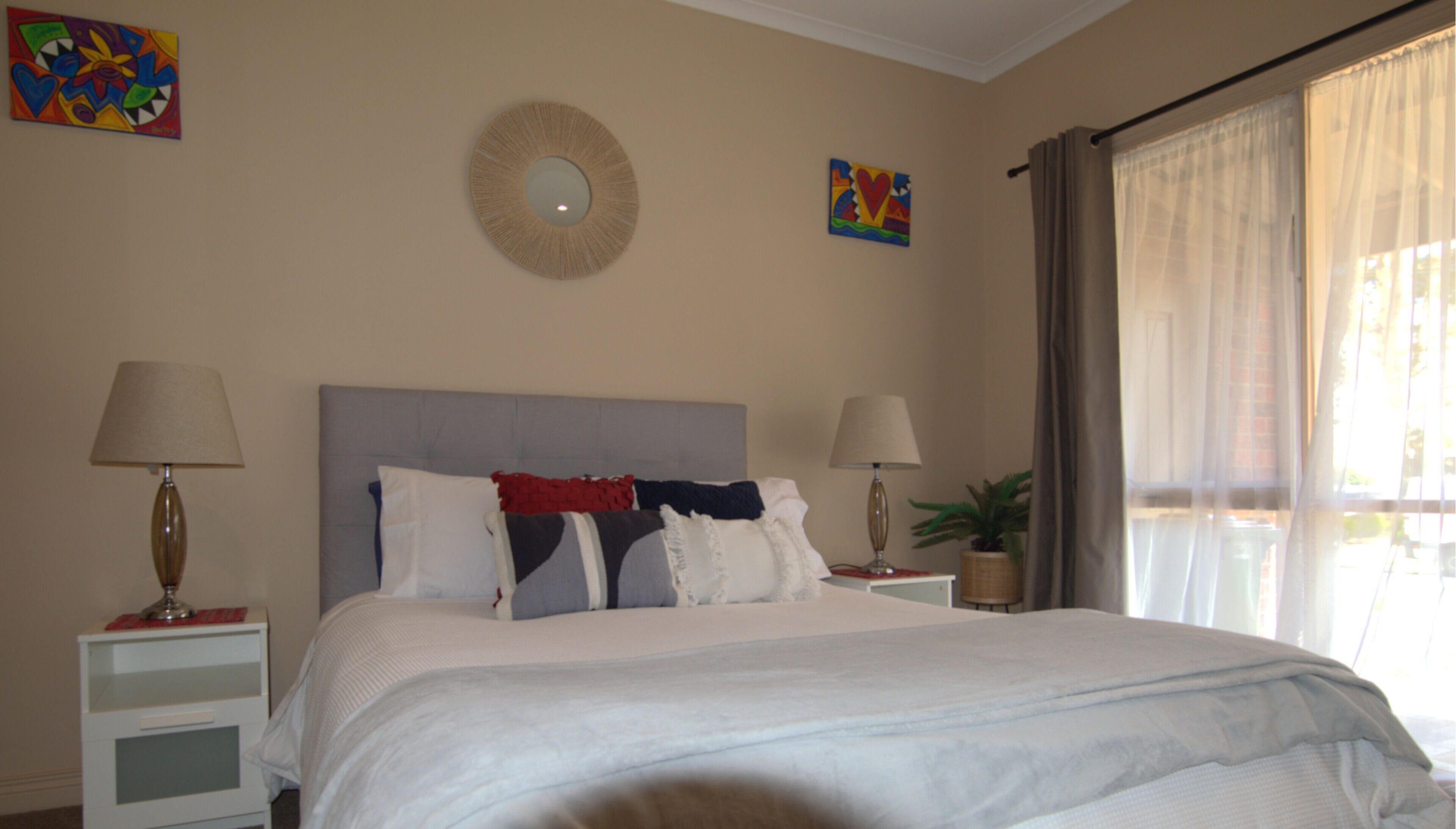 A neatly made bed with white bedding and multiple pillows, two bedside tables with lamps, a round mirror on the wall, and two colorful paintings.