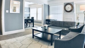 Ein Wohnzimmer mit Sofa, Couchtisch und Deckenventilator. Durch einen offenen Durchgang ist ein Essbereich mit Tisch und Stühlen zu sehen.