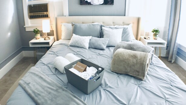 Ein ordentlich bezogenes Bett mit einer grauen Bettdecke, mehreren Kissen und einer grauen Schachtel auf dem Bett.