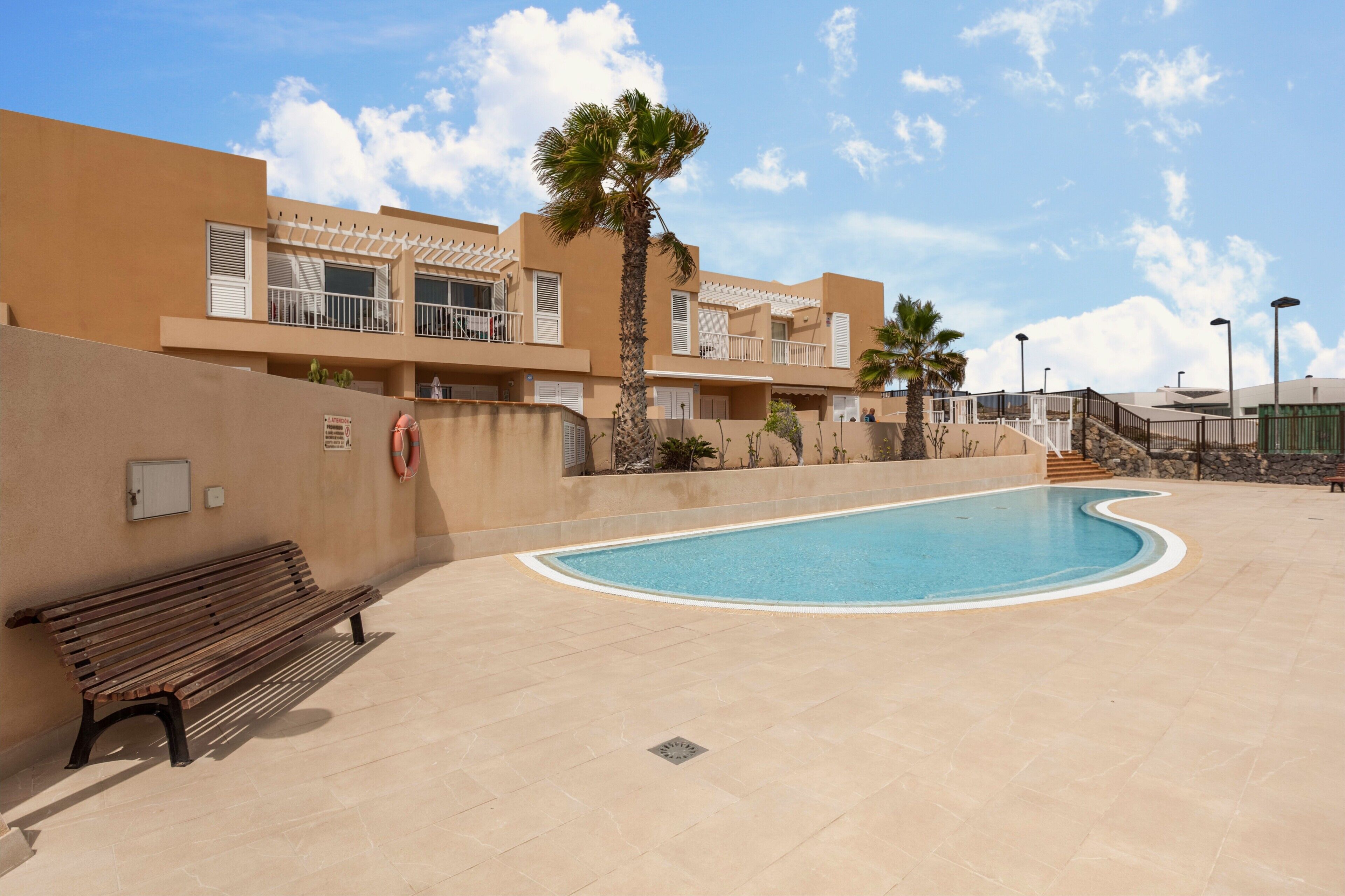 Zona de piscina con una banca, una palmera y un edificio con balcones.