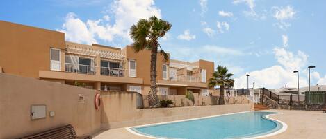 Zona de piscina con una banca, una palmera y un edificio con balcones.
