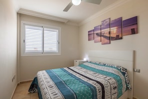 Een slaapkamer met een bed, een raam met jaloezieën, een plafondventilator en een muur met drie schilderijen.