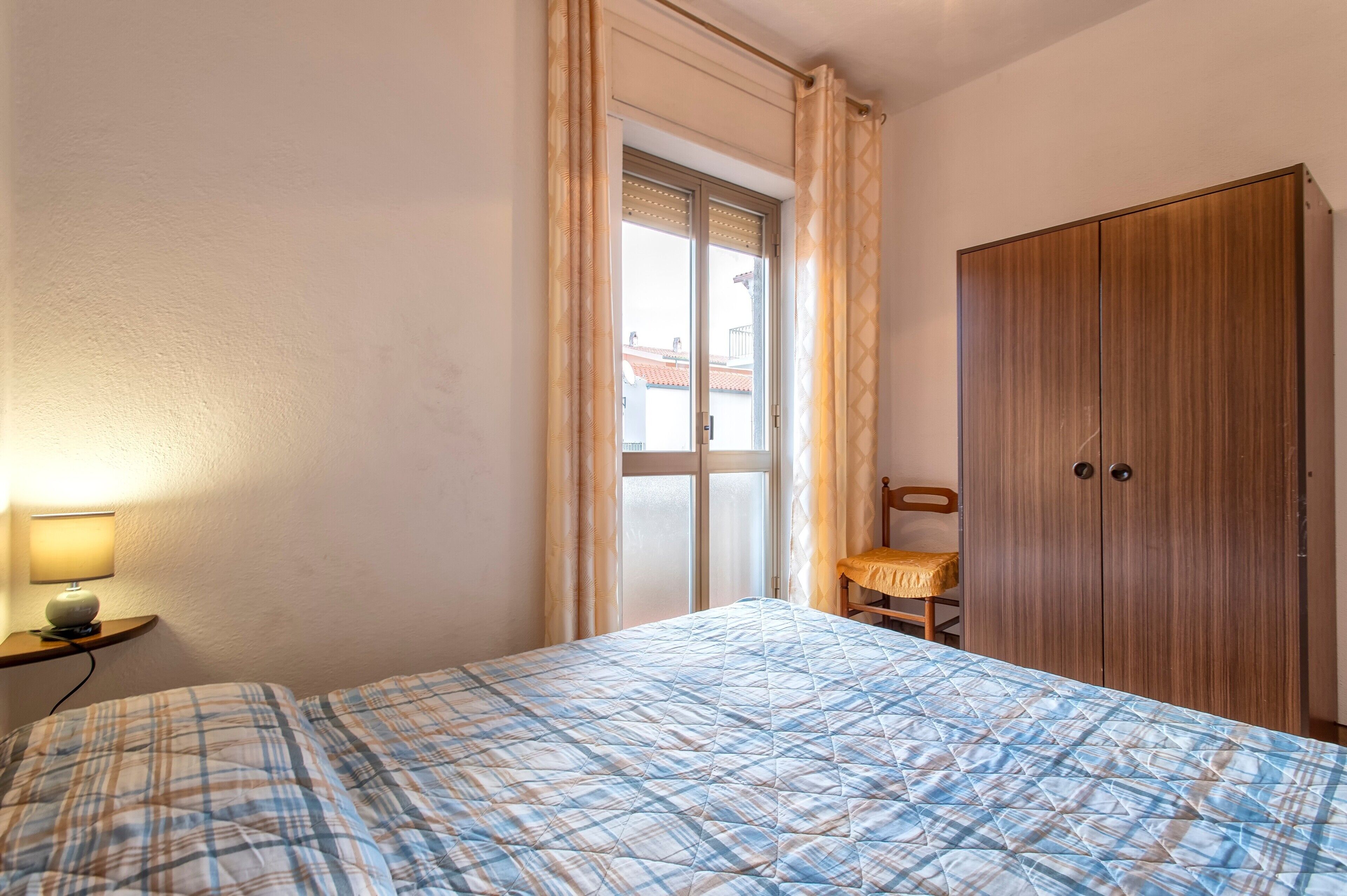 A bedroom with a bed, a wooden wardrobe, a chair, a nightstand with a lamp, and a window with curtains.