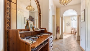 A wooden bench with magazines, a mirror, and a chandelier in a hallway with tiled flooring and columns. - Holiday Apartment 'Casa Zanoni' with Lake View, Shared Garden, Balcony & Wi-Fi (Arco)