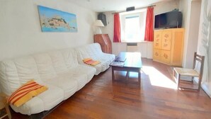 Un salon avec un canapé blanc, une table basse en bois, un téléviseur posé sur un meuble, un buffet orné de motifs floraux et une fenêtre avec des rideaux rouges.