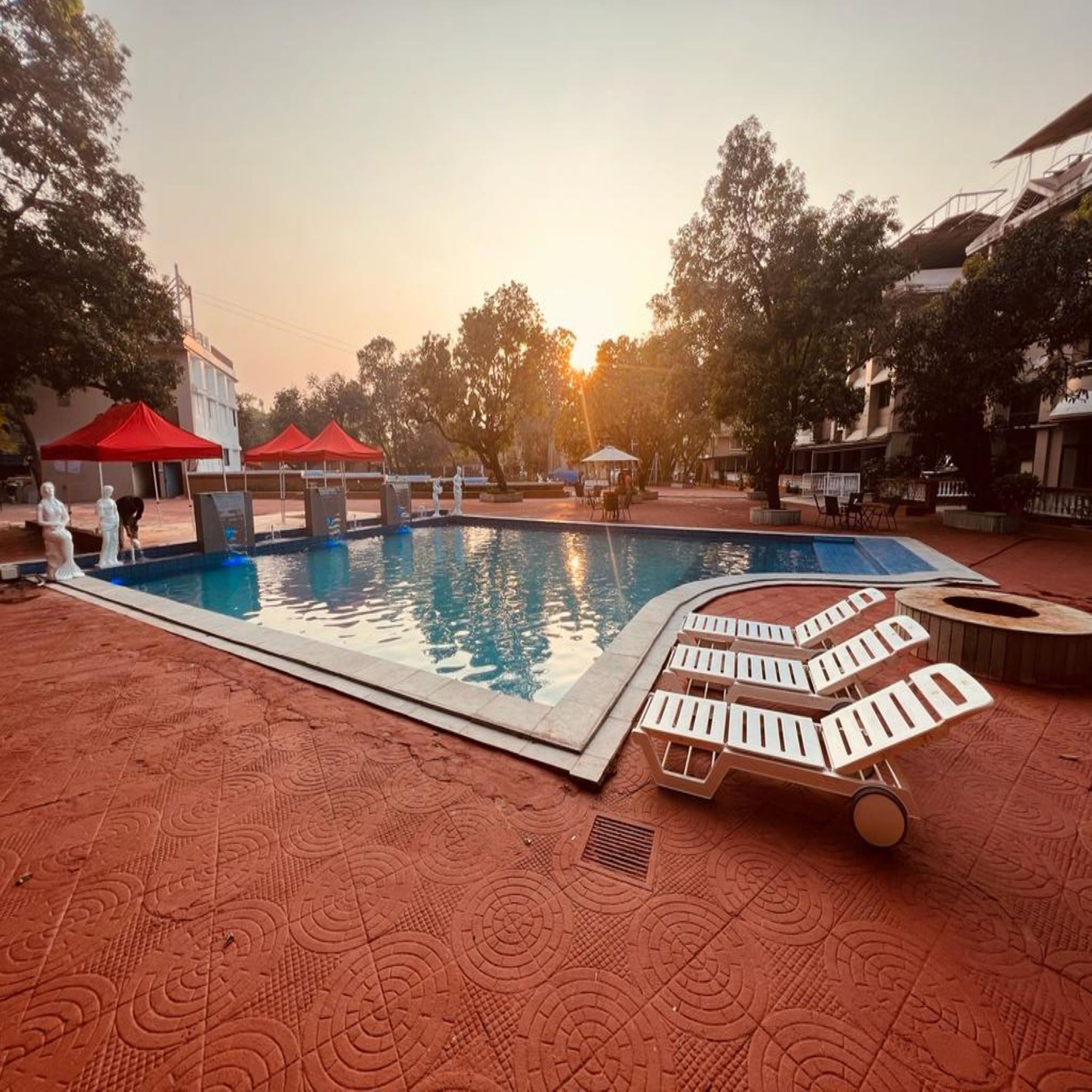 A swimming pool with lounge chairs and red canopies, surrounded by trees and buildings.