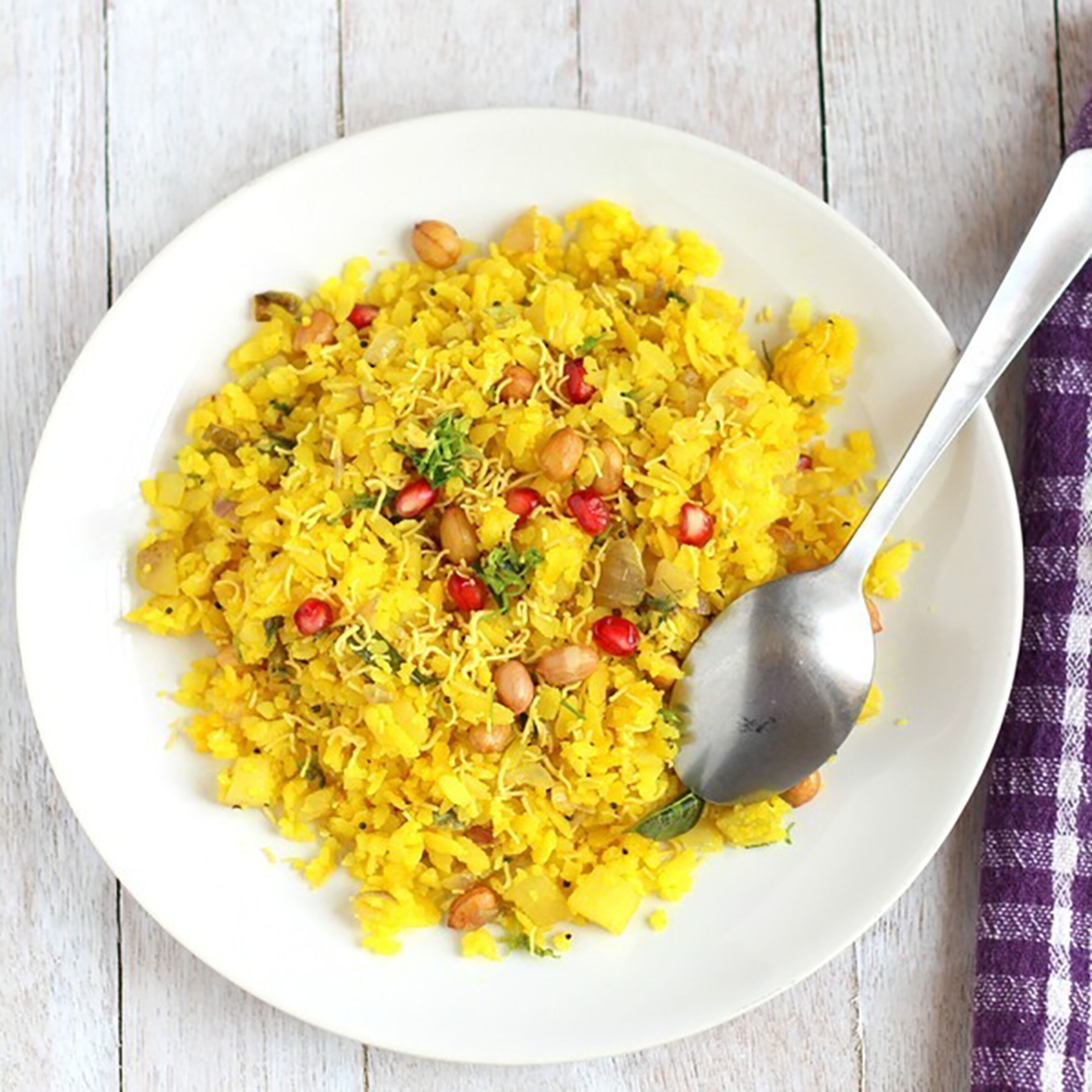 A white plate of yellow rice with peanuts and pomegranate seeds, accompanied by a spoon.