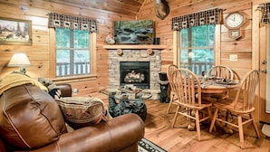 L’intérieur chaleureux d’un chalet avec une cheminée en pierre, des murs en bois, une tête de cerf accrochée au mur et un coin repas avec des chaises en bois et une table basse en verre.