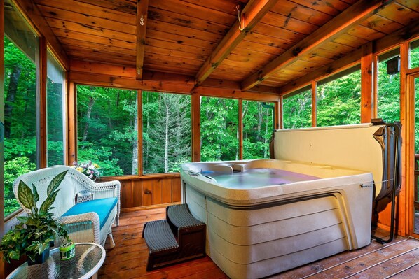 A wooden deck with a hot tub, wicker furniture, and large windows overlooking a forest. - Beautiful cabin! Book now! 2 master suites - Sleeps 6 - Hot Tub  (Sevierville)
