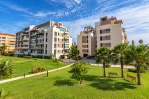 Een modern appartementencomplex met meerdere verdiepingen, balkons en een verzorgd gazon met palmbomen en een bankje.
