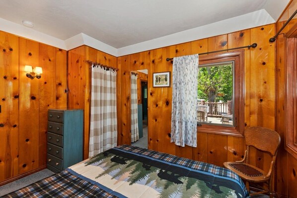 Una habitación con paneles de madera, una cama con colcha estampada, una silla, un aparador y una ventana con cortinas.