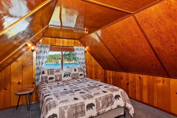 Un dormitorio acogedor en el ático, con techo inclinado, una cama con colcha estampada, un taburete pequeño y vista a una piscina exterior.
