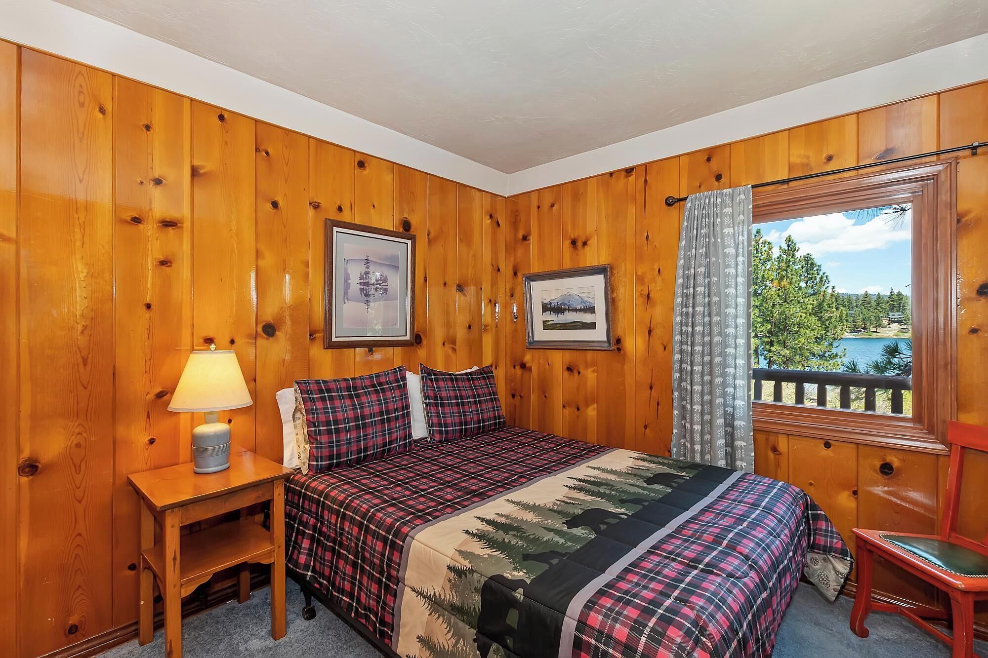 A wooden-paneled room with a bed, a nightstand, a lamp, and a window with a view of trees and a lake.