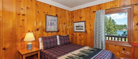 A wooden-paneled room with a bed, a nightstand, a lamp, and a window with a view of trees and a lake.