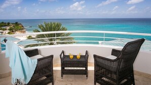 Un balcon agrémenté de meubles en osier et d’une table où sont disposées des boissons, offrant une vue sur un océan turquoise et une plage.