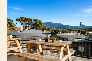 A rooftop patio with wooden tables and chairs, a coffee maker, and a view of a residential area with houses and mountains in the background.