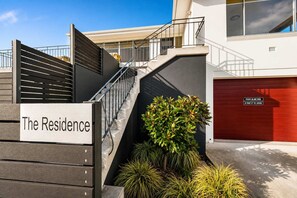 A residential property with a sign reading 'The Residence,' a red garage door, a staircase leading to a second floor, and a small garden area with plants. - "The Residence" Sleeps 11. 10 min to Airport/CBD. (Warrane)