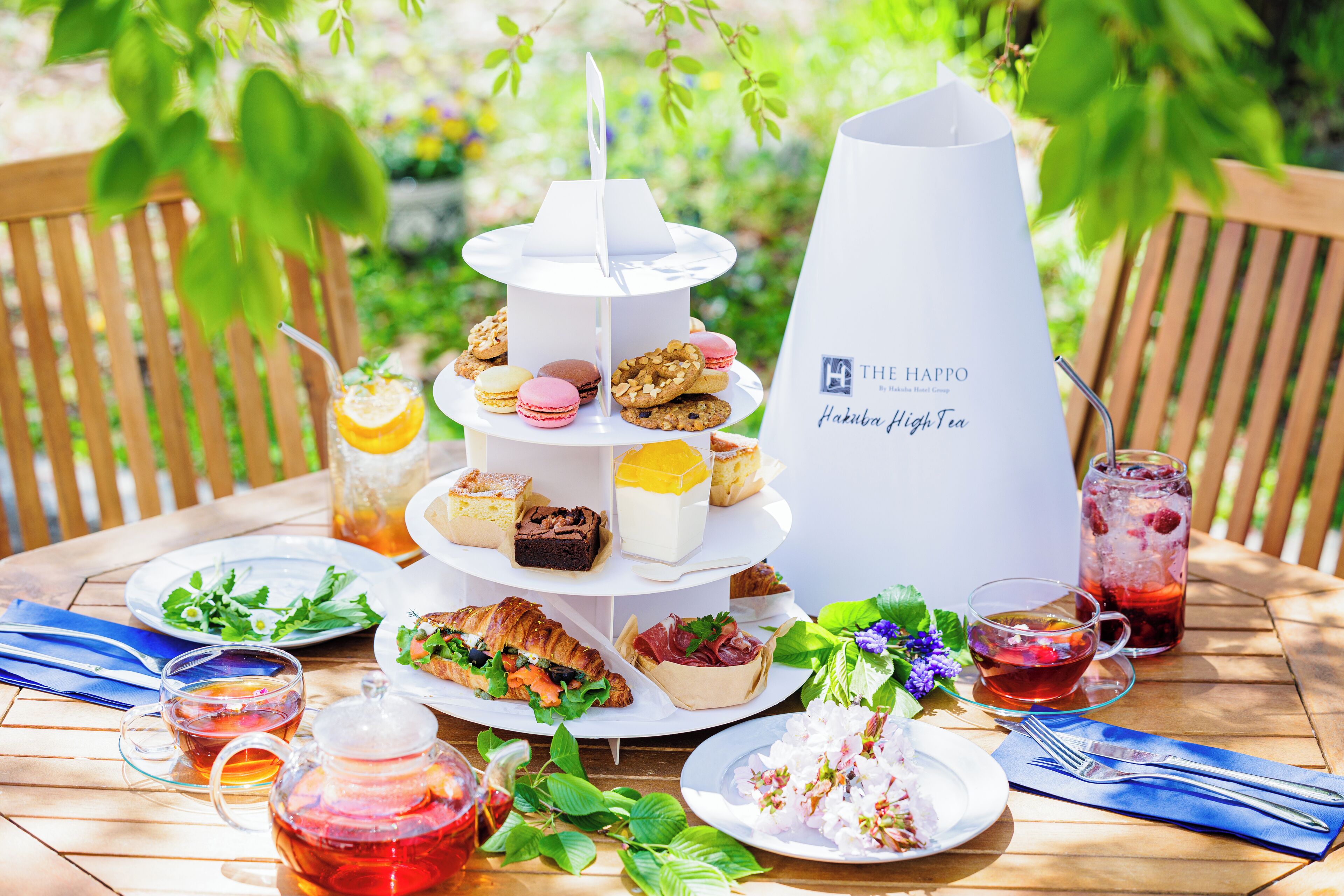 Suporte de sobremesas escalonado com diversos doces e um jarro de bebidas rotulado como "The Happo Pakuba High Tea" sobre uma mesa de madeira ao ar livre.