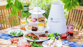 Suporte de sobremesas escalonado com diversos doces e um jarro de bebidas rotulado como "The Happo Pakuba High Tea" sobre uma mesa de madeira ao ar livre.