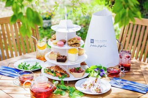 Suporte de sobremesas escalonado com diversos doces e um jarro de bebidas rotulado como "The Happo Pakuba High Tea" sobre uma mesa de madeira ao ar livre.
