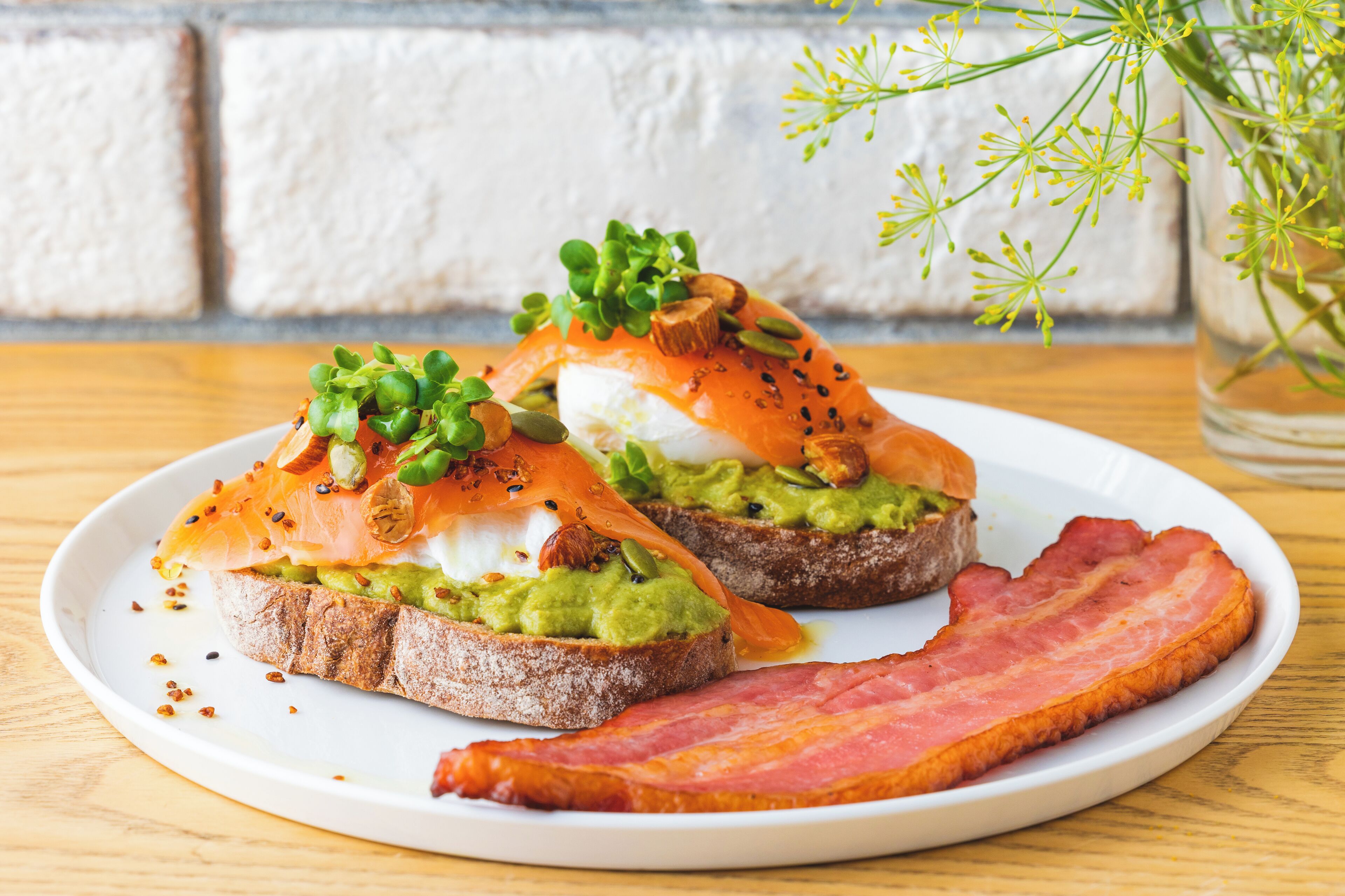 Une assiette avec deux sandwichs ouverts, l’un garni d’avocat et l’autre de saumon fumé, le tout agrémenté d’herbes fraîches et de graines, et une tranche de bacon à côté.