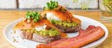 Une assiette avec deux sandwichs ouverts, l’un garni d’avocat et l’autre de saumon fumé, le tout agrémenté d’herbes fraîches et de graines, et une tranche de bacon à côté.
