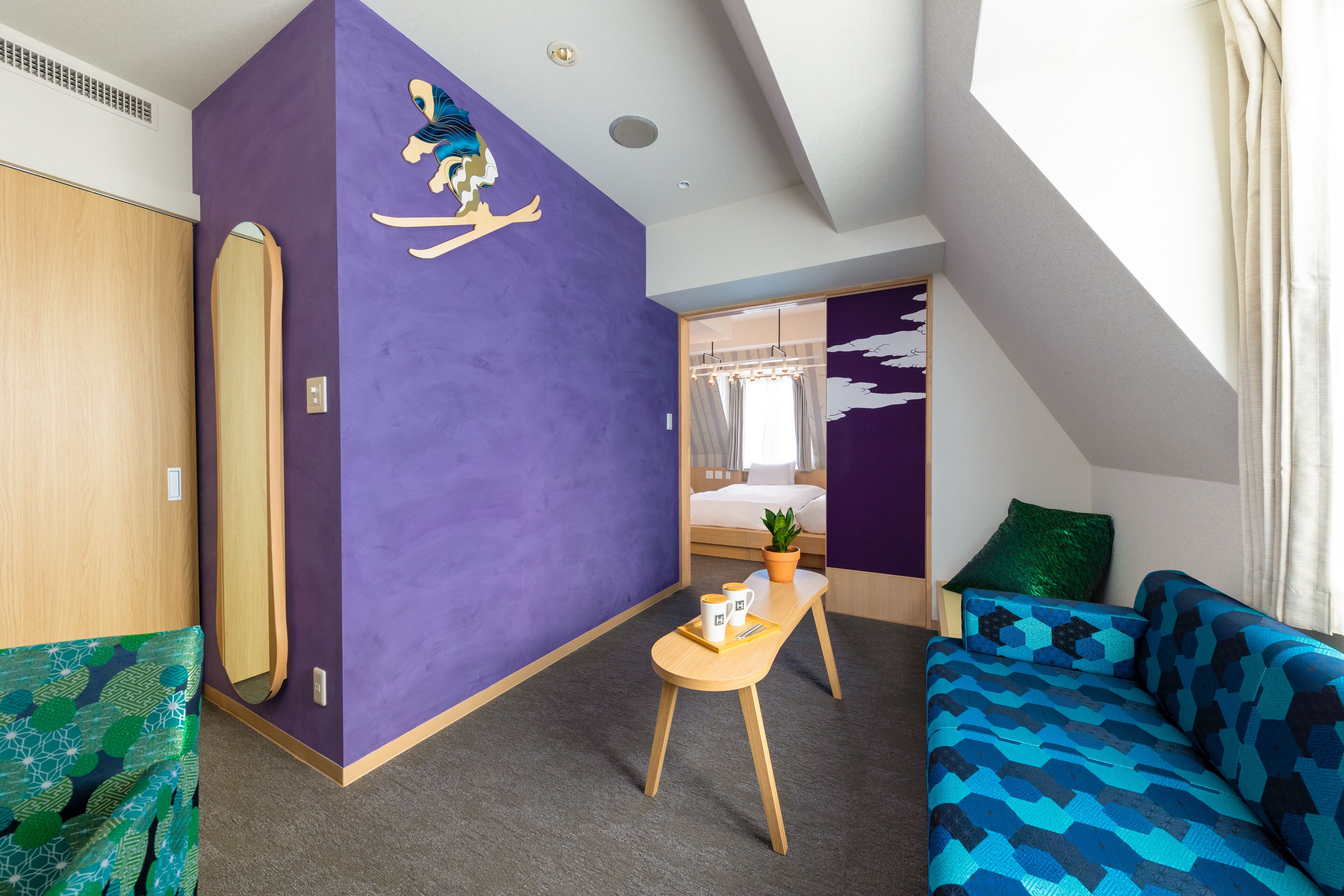 A modern room with a purple accent wall, a wooden wardrobe, a blue patterned sofa, a small wooden table with a plant, and a bed with a green pillow.