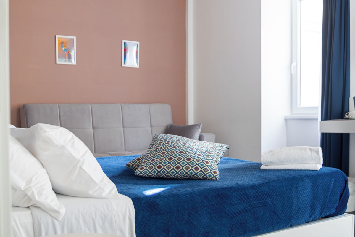 A bedroom with a bed, a headboard, a window with curtains, and exposed wooden beams on the ceiling.