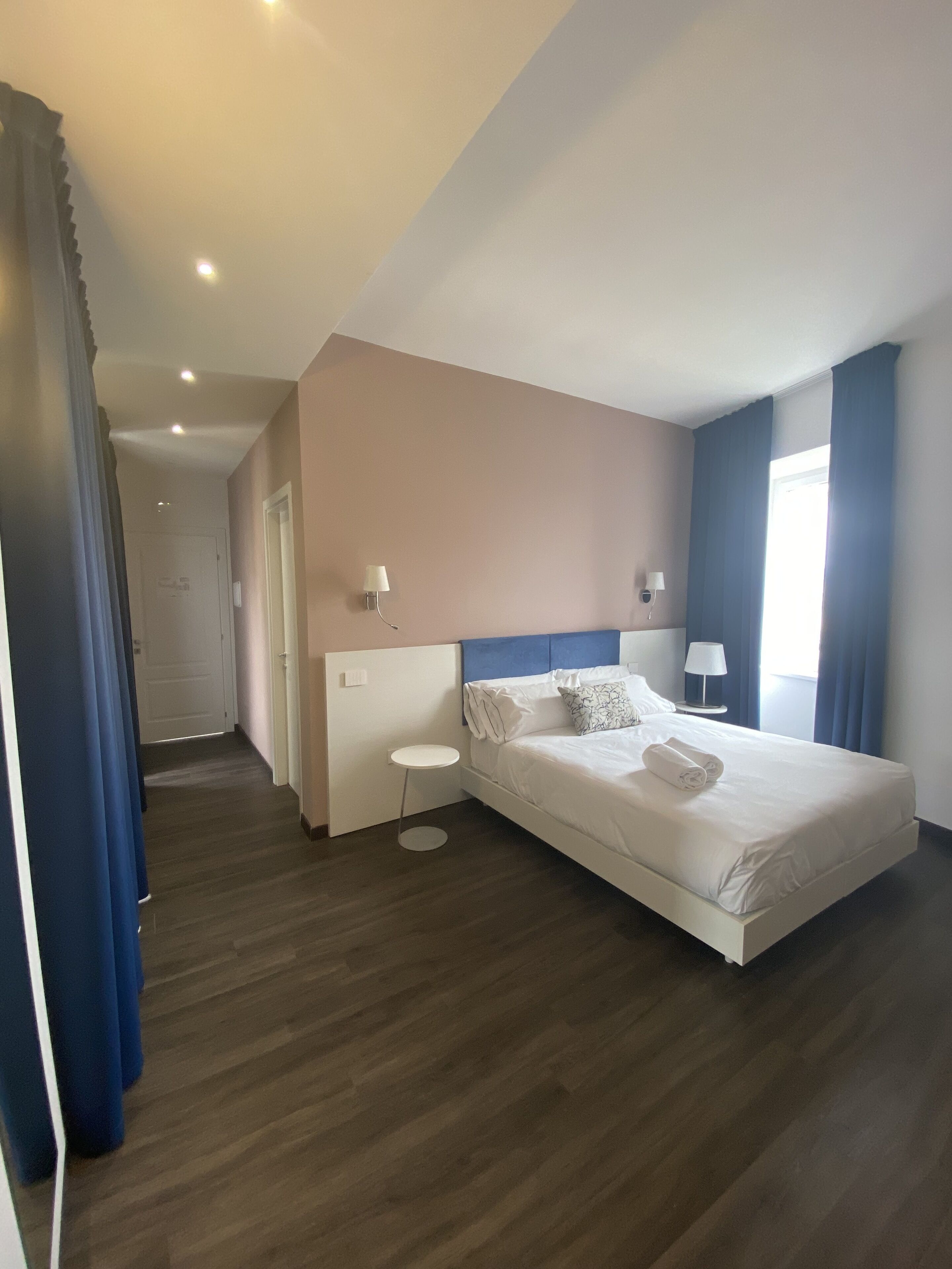 Quarto de hotel com uma cama grande, uma mesa de cabeceira pequena, uma luminária na parede e uma porta que dá para outro cômodo.