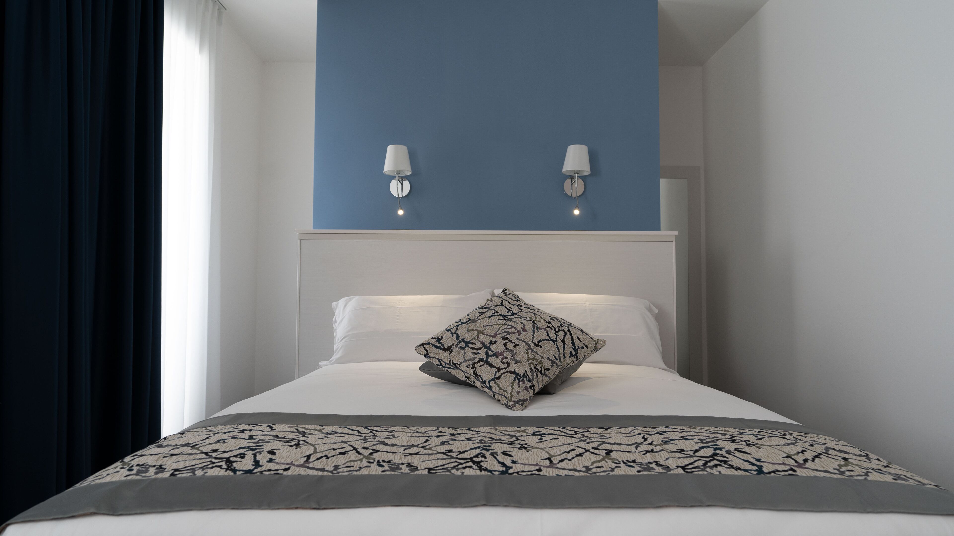 Una cama bien tendida con una colcha estampada y un cojín decorativo, un cabecero blanco y dos lámparas fijadas a la pared.