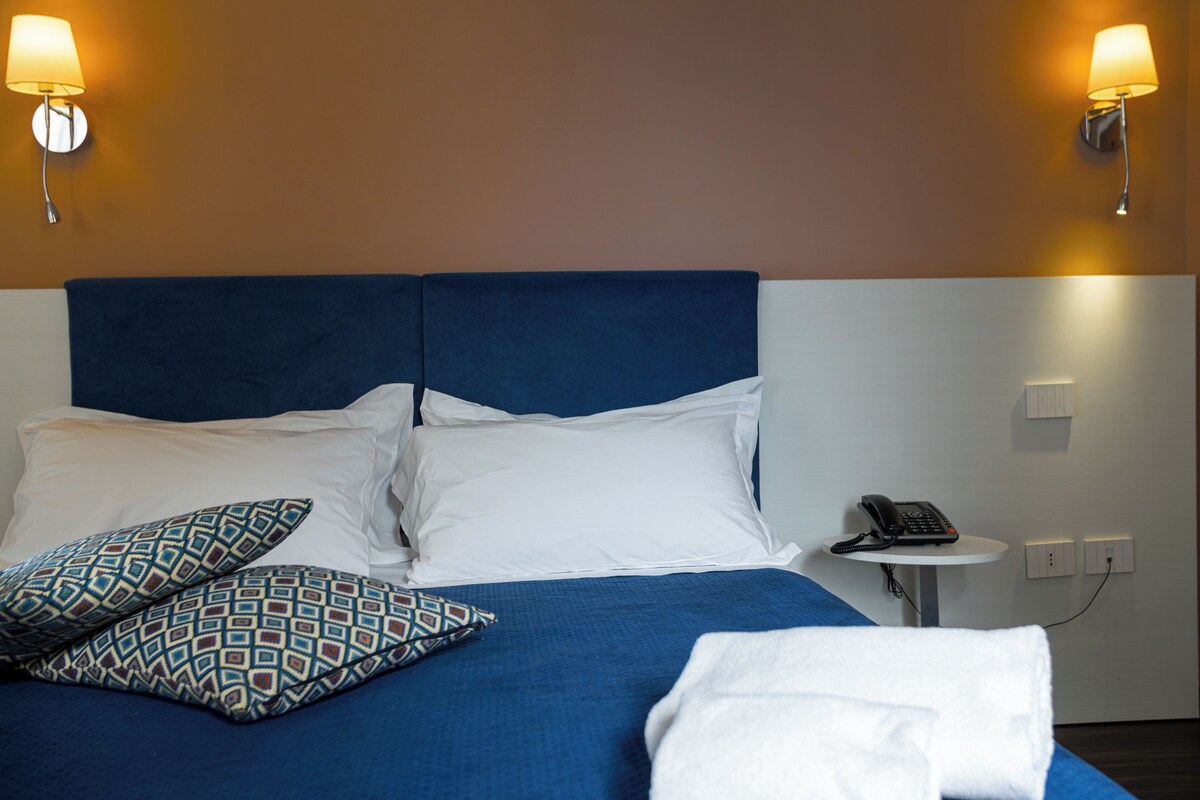 A hotel room with a blue bedspread, white pillows, patterned pillows, a white bathrobe, and two wall-mounted lamps.