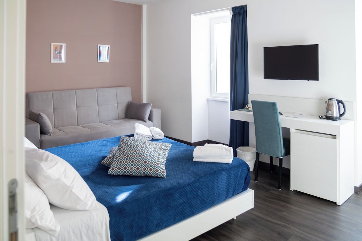 A modern hotel room with a bed, a desk, a chair, a TV, and a window with curtains.