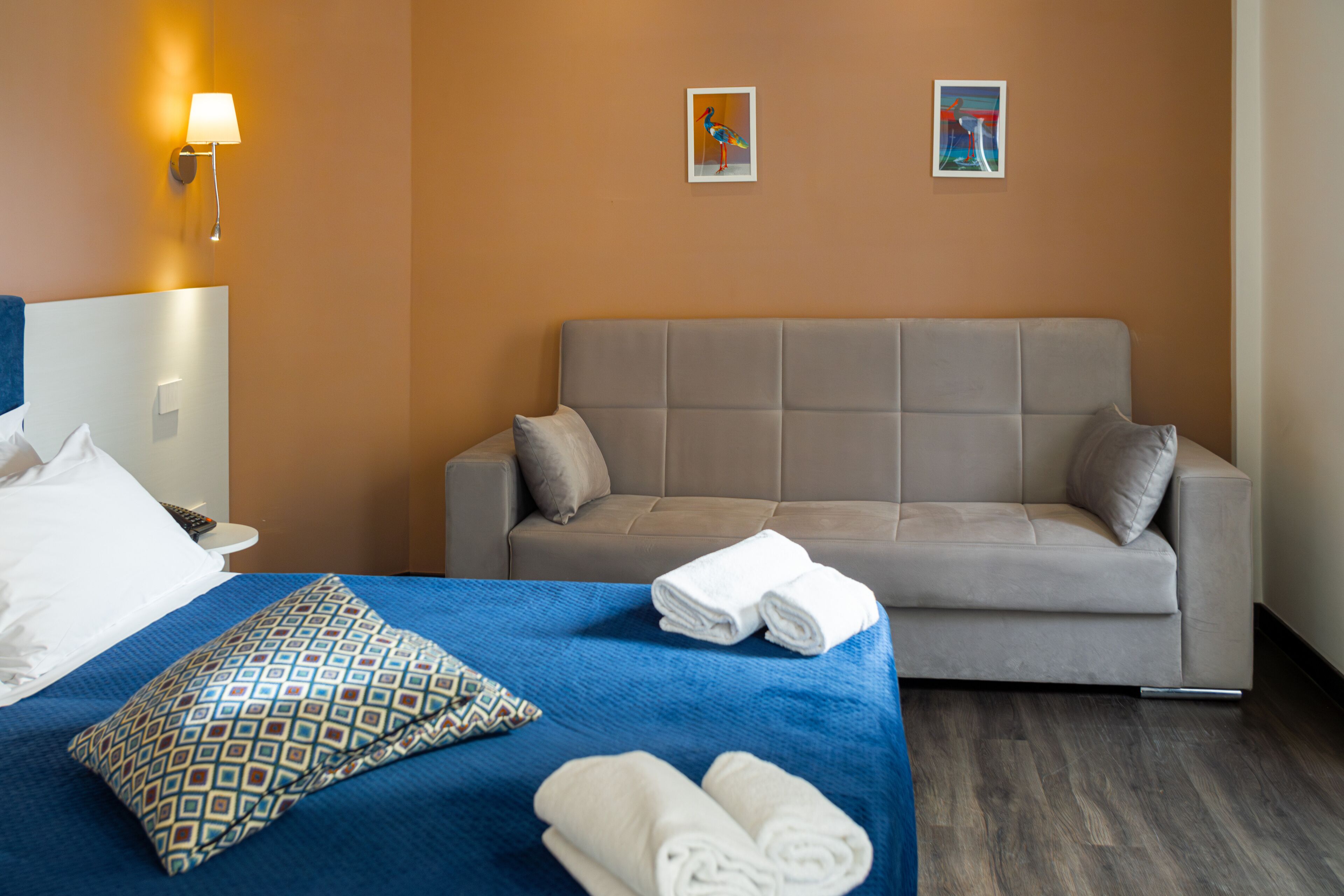 A hotel room with a bed, a sofa, a patterned pillow, and two artworks on the wall.
