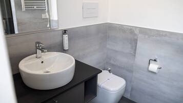 Un baño moderno con lavamanos blanco, encimera oscura y paredes cubiertas de baldosas grises.