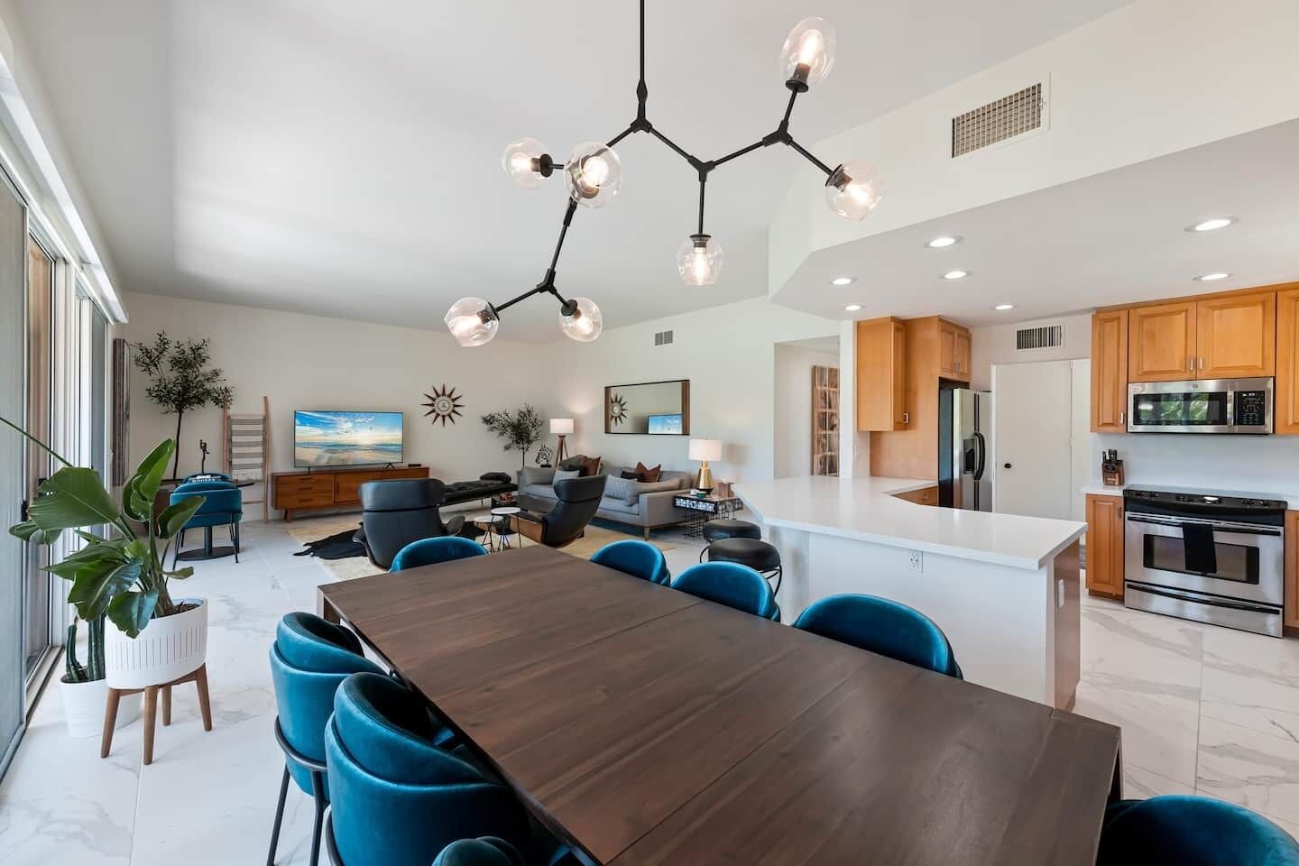 A modern kitchen with stainless steel appliances, a dining area with a wooden table and blue chairs, and a living area with a sofa and a TV.